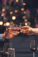 Cheers to Success: Two hands clinking glasses of amber liquor, a toast to celebration and camaraderie in a dimly lit, festive setting.