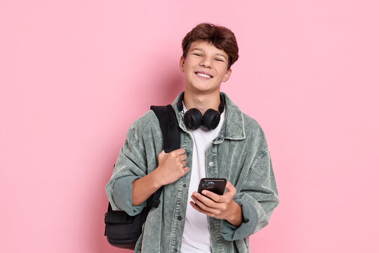 Happy teenage boy with headphones, smartphone and backpack on pink background