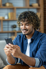 Young man at home using an app on his phone, sitting on a sofa in the living room holding a phone...