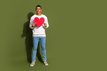 Young man in white sweater holding red heart on khaki background, expressing joy and positivity, perfect for promoting love