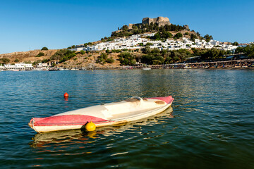 Old kayak boat in shallows of sandy beach in Bay of Lindos, with Lindos town and  Acropolis in background (Rhodes, Greece)