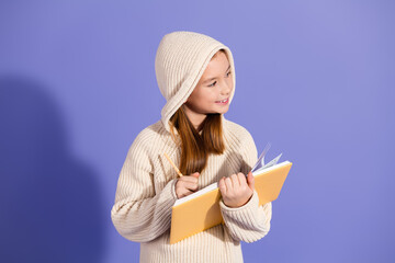 Young girl holding a notebook and pencil against a purple background, wearing a hoodie sweater,...