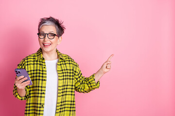 Portrait of pretty aged lady hold phone point finger empty space plaid shirt isolated on pink color background