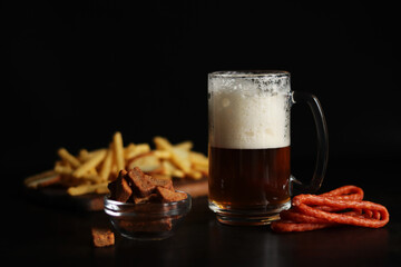 Glass with light beer and snacks on a dark background. Beer, French fries and hunting sausages. Glass on a wooden board and snacks. Light beer and bread croutons. Traditional glass beer mug