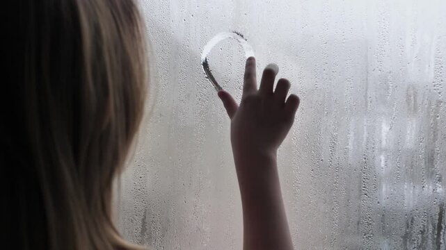 little teenage girl uses finger to draw heart shape on wet foggy window glass. Condensation on windows used by child to draw heart. Expression of love. Child love