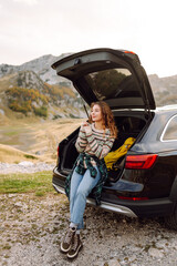 Naklejka premium Happy woman sits in the trunk of her car, parked on the side of a winding road that cuts through the rocky terrain. Lifestyle, travel, tourism, nature, active life