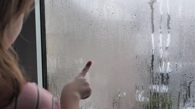 Red-haired child girl draws on wet window glass the shape of animal paw prints using only her hands and fingers. Drawings on wet windows. Rainy weather conditions. Condensation on windows