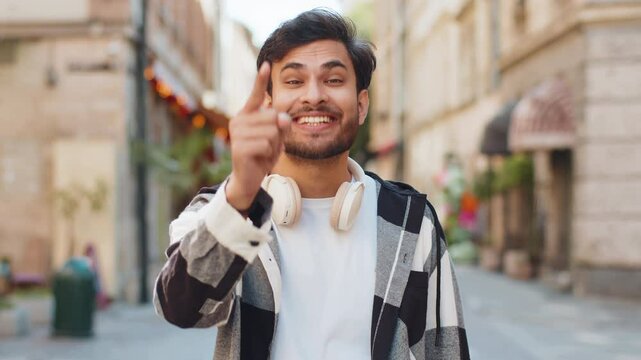 Hey you. Young Indian man tourist smiling excitedly and pointing to camera, choosing lucky happy winner, indicating to awesome you. Handsome Arabian guy standing in urban city street. Lifestyles.