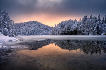 Fototapeta premium Frozen lake reflecting warm hues of a winter sunset, snow-covered trees and gently rolling hills. Winter colorful landscape with lake, snowy forest, golden sunset. Fairytale in Plitvice Lakes, Croatia