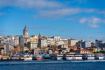 Fototapeta premium The Galata Tower rises high above the rooftops of Istanbul with tourist boats on the banks of the Bosphorus in the foreground