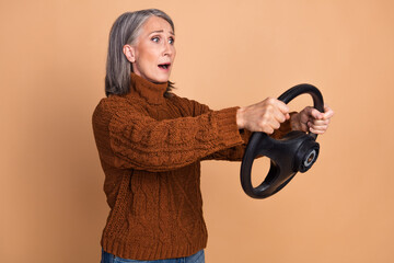 Obraz premium Surprised Elderly Woman in Brown Sweater Holding Steering Wheel Against Beige Background