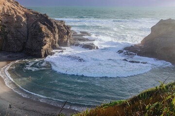 Powerful waves crash against rocky cove; people swim in the surf. Piha, Auckland, New Zealand
