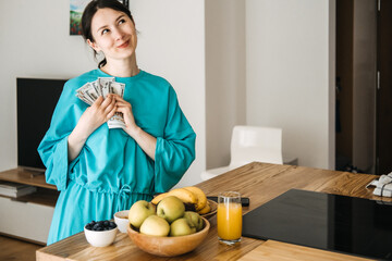 Smiling woman holding money in cozy kitchen with fresh fruits. Financial wellness, budgeting habits, healthy lifestyle, and abundance mindset inspiration.