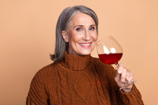 Elegant senior woman enjoying a glass of wine against a beige background, showcasing sophistication and joyful mood in cozy autumn fashion.
