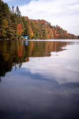 Tranquil lake scene in autumn with a white an blue Canadian hut situated on the shore, next to a small dock. background with trees in vibrant autumn colors and beautiful lake reflection