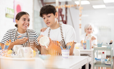 Happy young guy and girl spending quality time together in pottery studio, creating and decorating ceramic cups. Creativity and bonding concept..