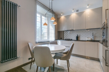 The interior of the kitchen in pastel colors. Kitchen set with elegant design and stylish accessories. Dining table with upholstered chairs. A large metal heating battery on the wall.
