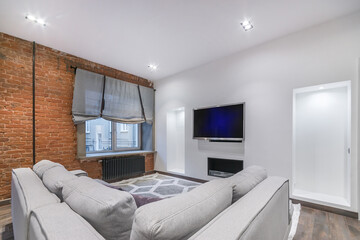 Modern living room interior with loft-style decor elements. The wall is made of red brick. A light gray sofa and a large TV on the wall.