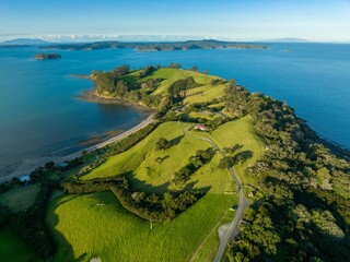 Aerial view of a lush green coastal farm, with a small house nestled amongst rolling hills....