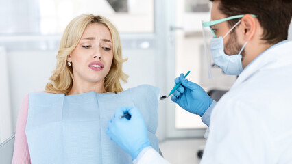 Scared young blonde woman female patient sufffering from dental phobia, sitting at dental chair, looking at tools in dentist hands with frightened face expression, avoid treatment at dentistry