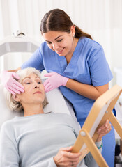 Obraz premium Pleasant young woman doctor consulting aged woman patient lying on clinical chair about face aesthetic