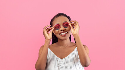 Joyful young black lady in stylish summer dress touching sunglasses and smiling at camera on pink studio background. Lovely millennial African American woman posing in trendy outfit