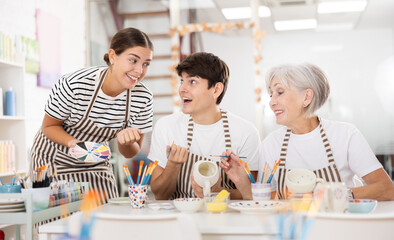 Enthusiastic senior female teacher sharing experience pottery craftsmanship, assisting happy young guy and girl, painting ceramic mugs and plates at table in workshop