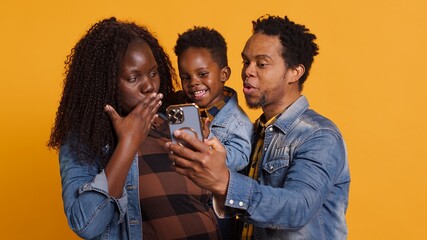 Cute happy family with a young kid take pictures on smartphone, carrying their son and sharing embrace in studio. African american parents feeling grateful for their toddler. Camera A.