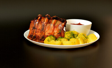 Large round plate of tasty lunch, young boiled potatoes with dill and baked pork ribs with tomato sauce on black background.