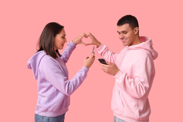 Young couple with mobile phones making heart with hands on pink background. Online dating