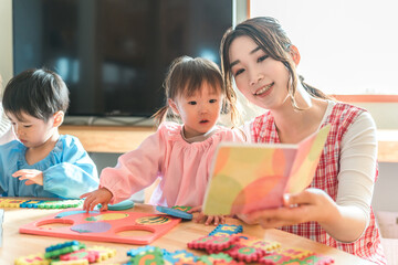 保育園・幼稚園・託児所・保育所で園児の女の子に絵本を読み聞かせする女性保育士・保母さん・シッター
