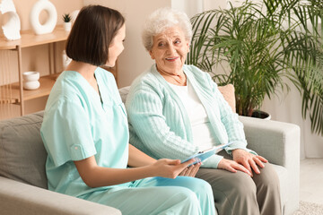 Young nurse with clipboard and senior woman sitting on sofa at home