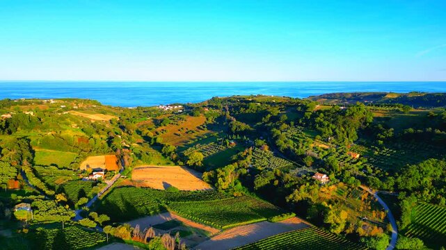 Smooth aerial push-in shot from Campofilone, swathes of green and brown fields lie on the Marche valleys and hills between roads, buildings, thrifty trees, vegetation, advancing into the Adriatic Sea