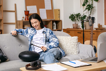 Beautiful young happy female composer with laptop and sticks playing glucophone on sofa at home
