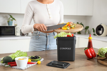 Young woman throwing vegetables into container for compost on table in kitchen