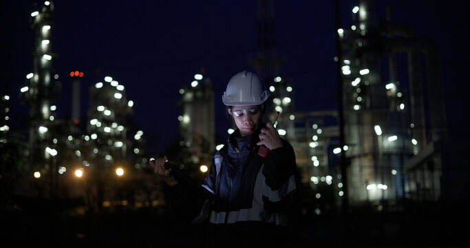 A female chemical  worker in safety gear uses a digital  tablet at a brightly lit refinery at night