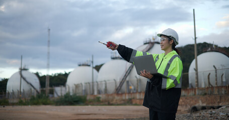 An industry engineer woman using a laptop with a white safety helmet standing in front of the oil...