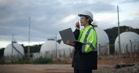 An industry engineer woman using a laptop with a white safety helmet standing in front of the oil...