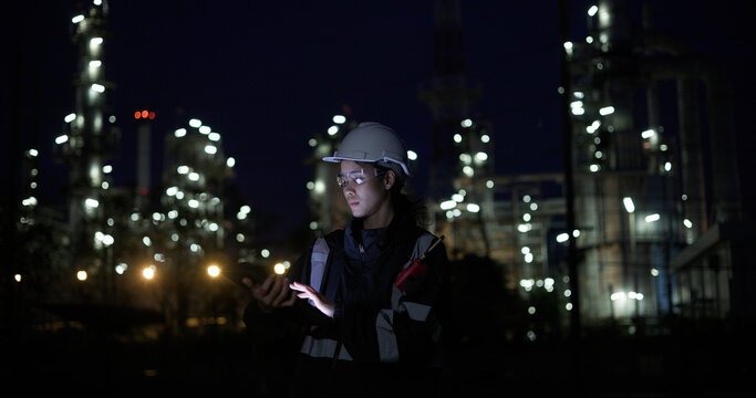 A female chemical  worker in safety gear uses a digital  tablet at a brightly lit refinery at night