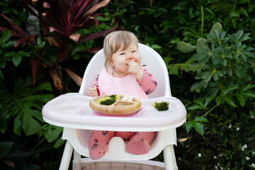 Cute Caucasian baby girl sitting in a high chair and eating healthy food. Baby led weaning concept. Baby eating finger foods in a tropical background, with copy space. Baby eating raspberry