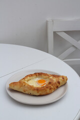 Vertical food photo. Fresh Adjarian style khachapuri with cheese and yellow yolk of an egg on round plate on white kitchen table near chair. Concept of lunch in cafe, national Georgian cuisine, eat