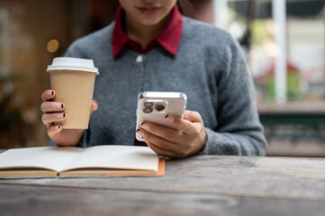 A woman reads text messages on her smartphone and enjoys a hot drink while sitting at a table.