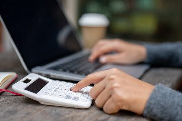 A close-up of a woman using a calculator while working on her laptop at a table outdoors.