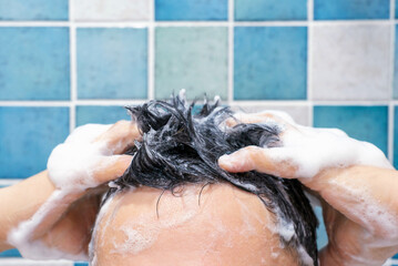 Male hands wash their hair with shampoo and foam against background of blue tiles, front view.