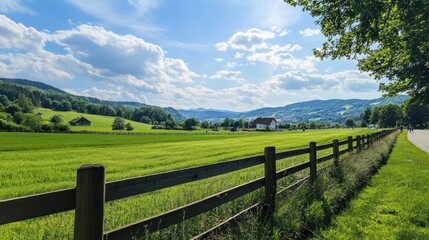 Fototapeta premium Sunny field with fence, distant houses and hills, road on side, suitable for travel or real estate