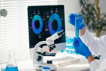 Scientist wearing blue gloves holding and examining test tube with blue liquid in laboratory with microscope and computer displaying charts in background