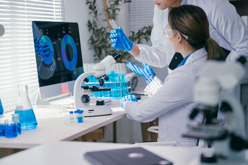 Two medical researchers in white coats and blue gloves are analyzing data on a computer screen while holding a test tube and discussing their findings in a modern laboratory