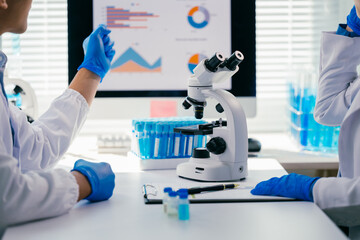 Two medical researchers wearing lab coats and gloves are discussing a medical research project in a laboratory, using a microscope and a computer displaying graphs and charts