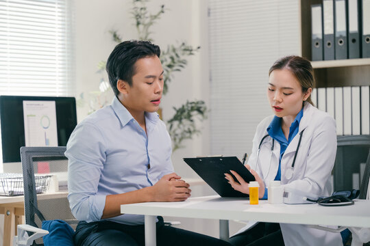 Female doctor taking notes on a clipboard while attentively listening to a male patient discussing his symptoms during a medical consultation in a hospital office setting