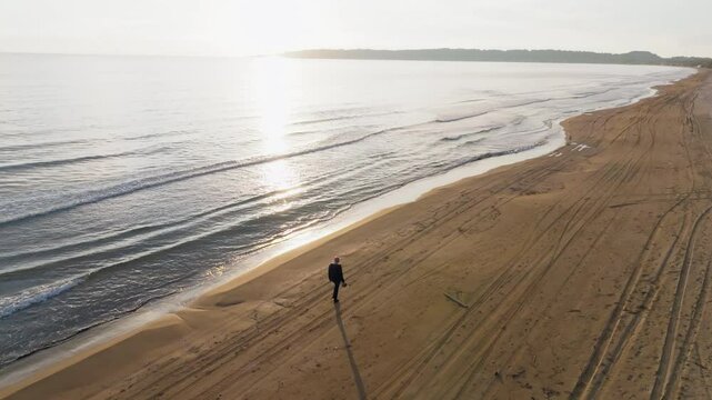 Slow orbit drone shot of a man walking on a sandy beach at sunset
